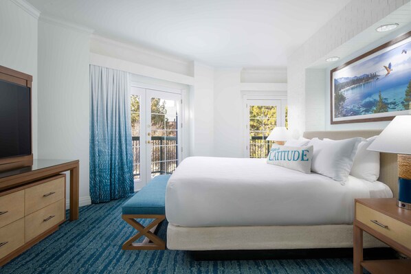 A hotel room with a large bed, a TV, a desk, and a balcony with curtains.