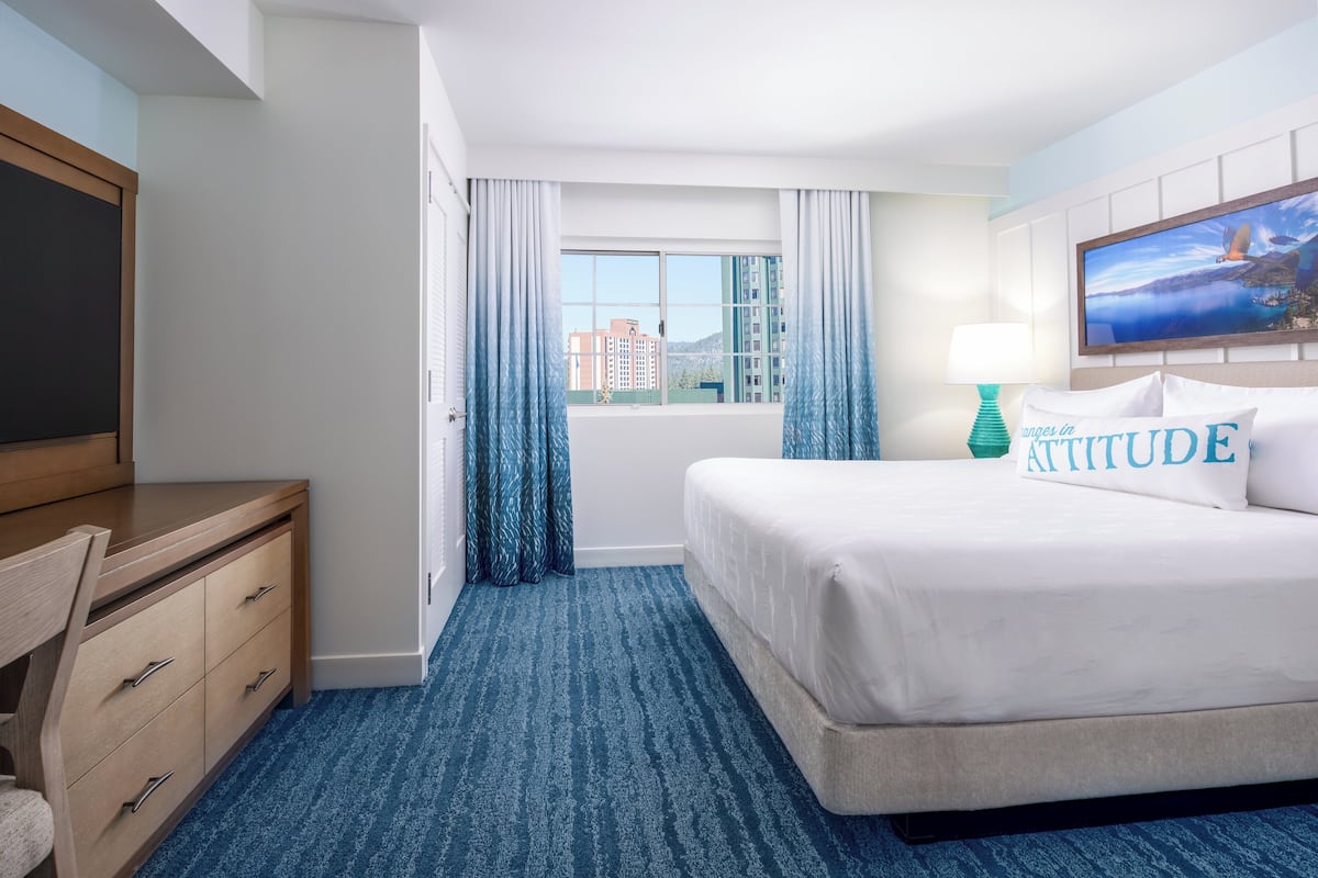 A hotel room with a bed, a desk, a chair, a window with curtains, and a view of buildings.