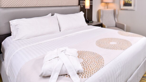 A neatly made bed with white linens and a towel, a headboard, a nightstand with a lamp, and a chair. - Holiday Inn Dallas-Richardson by IHG (Richardson)