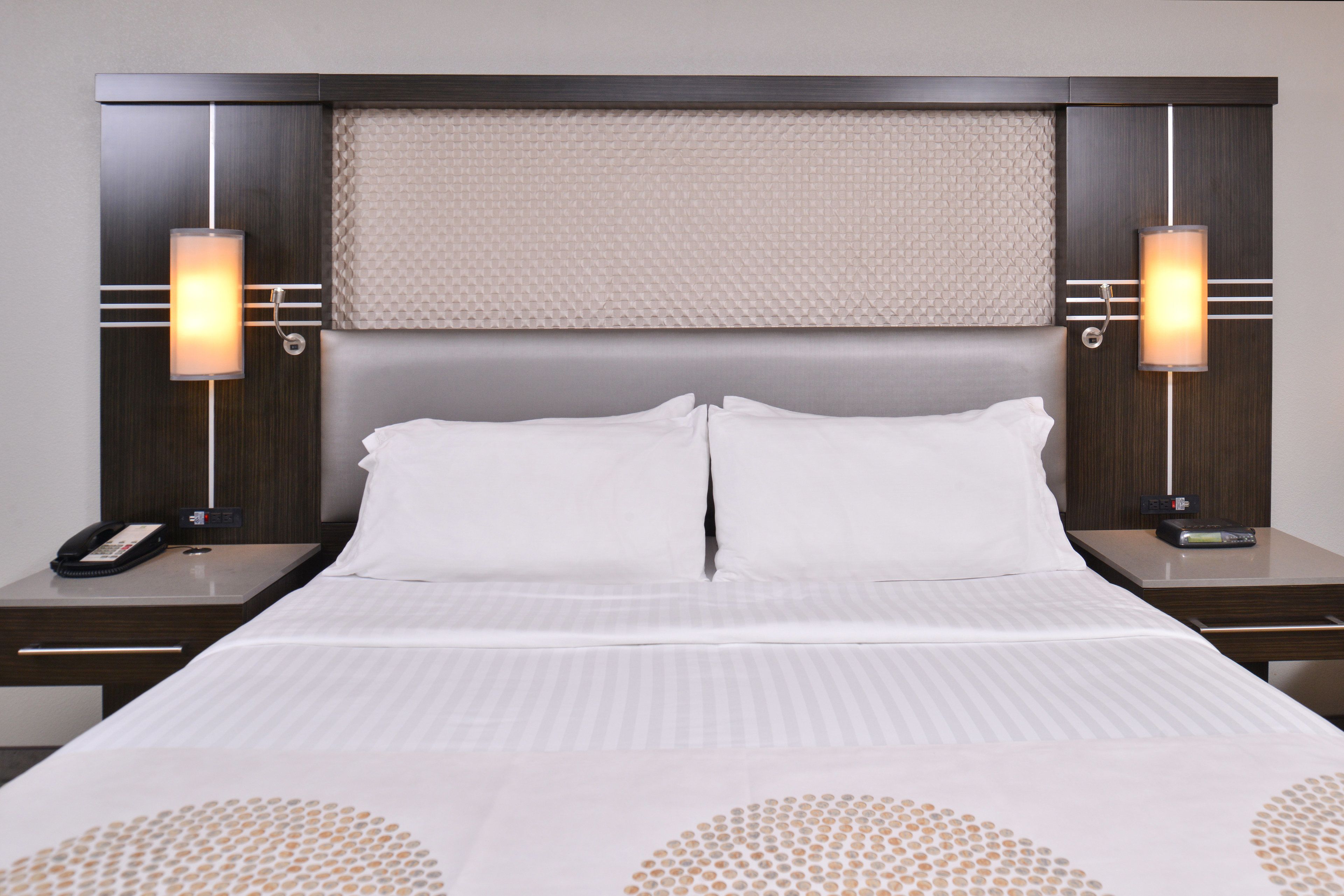 A hotel room with a large bed, two bedside tables, a headboard with a textured panel, and two wall-mounted lamps.