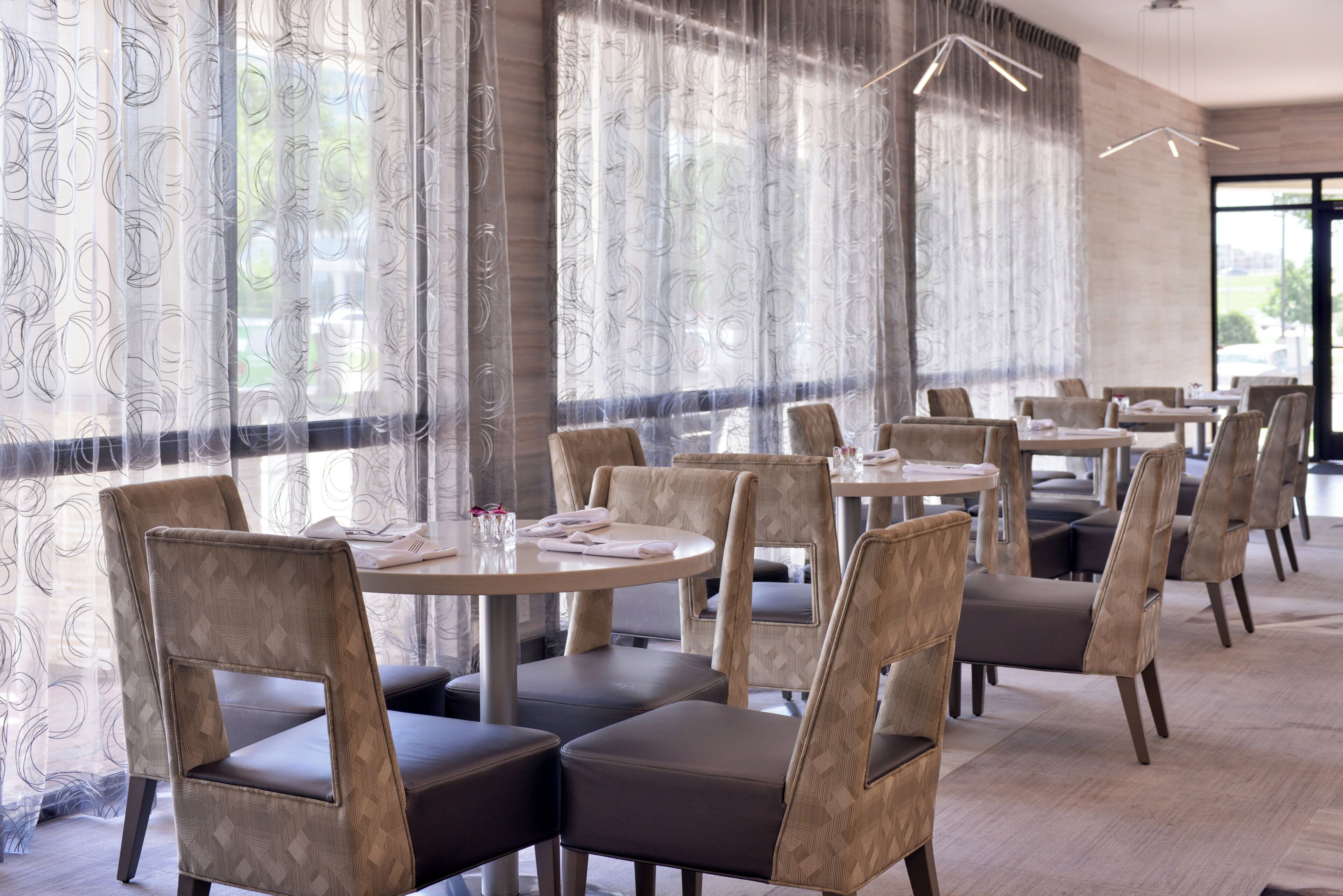 A restaurant interior with round tables and cushioned chairs, large windows with patterned curtains, and a modern chandelier.