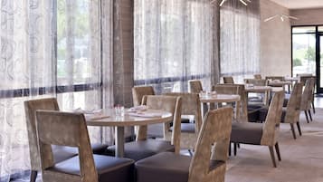 A restaurant interior with round tables and cushioned chairs, large windows with patterned curtains, and a modern chandelier.