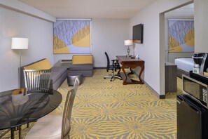 A hotel room with a glass dining table, a sofa with cushions, a desk with a lamp, a microwave, and a coffee maker.