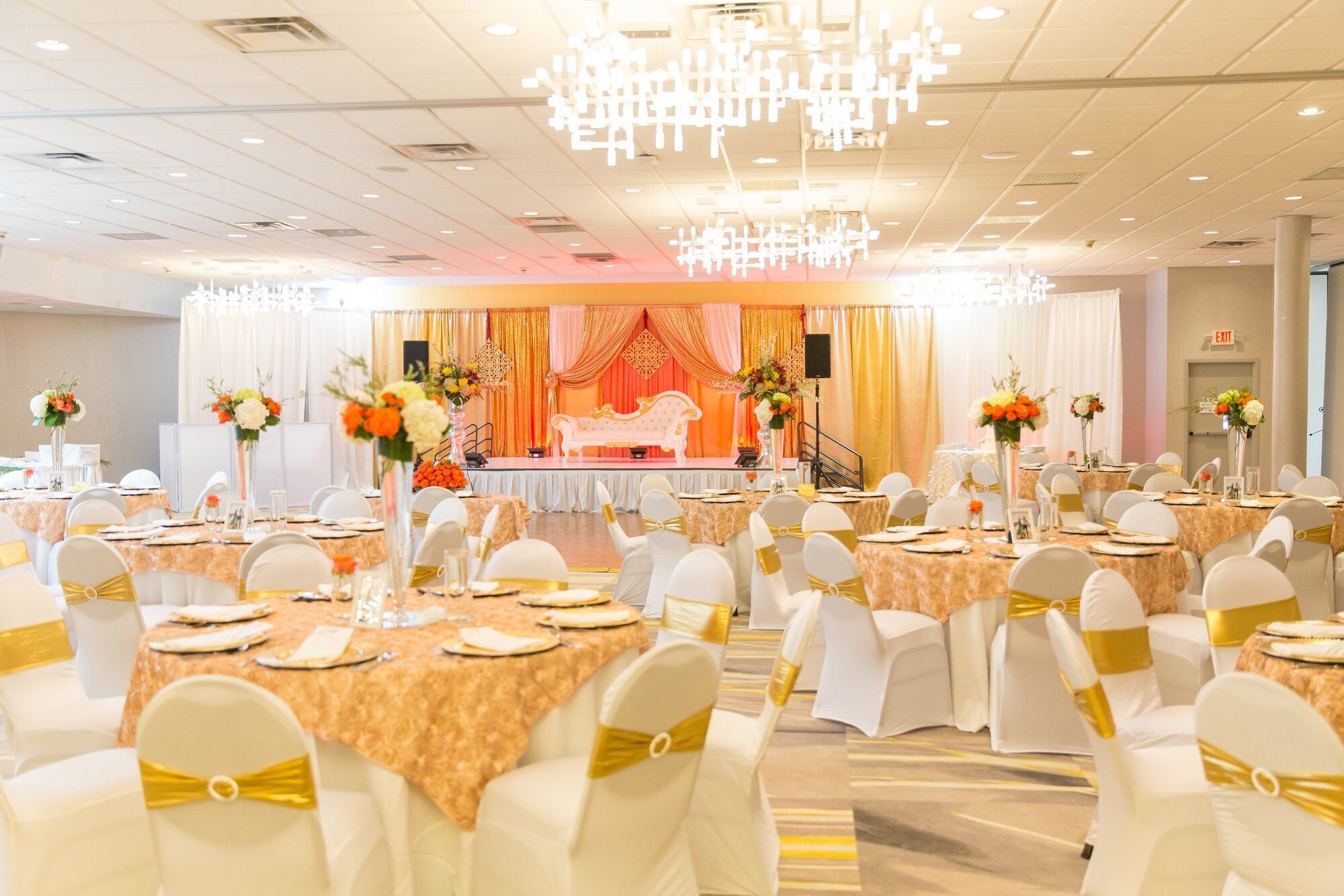 Een feestzaal met ronde tafels, witte stoelen met gouden linten en een podium met een rode achtergrond.