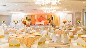 Un salón de banquetes con mesas redondas, sillas blancas con cintas doradas y un escenario con un telón de fondo rojo.