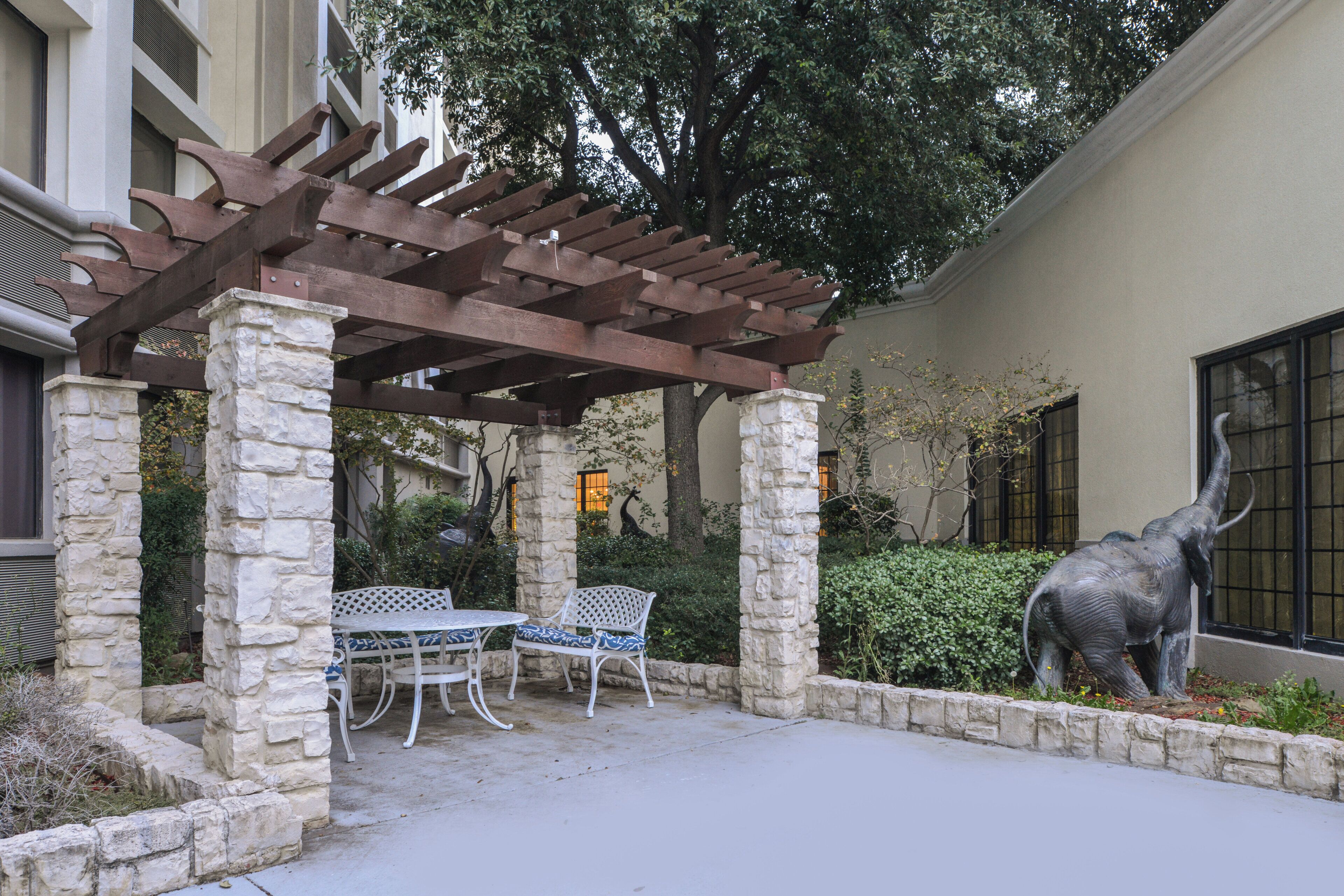 A covered patio area with stone pillars, a pergola, two white metal chairs, and a statue of an elephant.