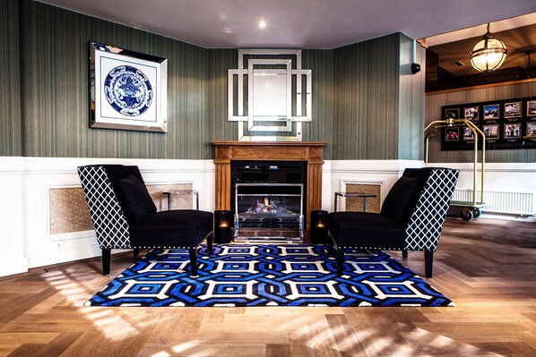 A room with a fireplace, two armchairs, a patterned rug, and a framed artwork on the wall. - Carlton Ambassador Hotel (The Hague)