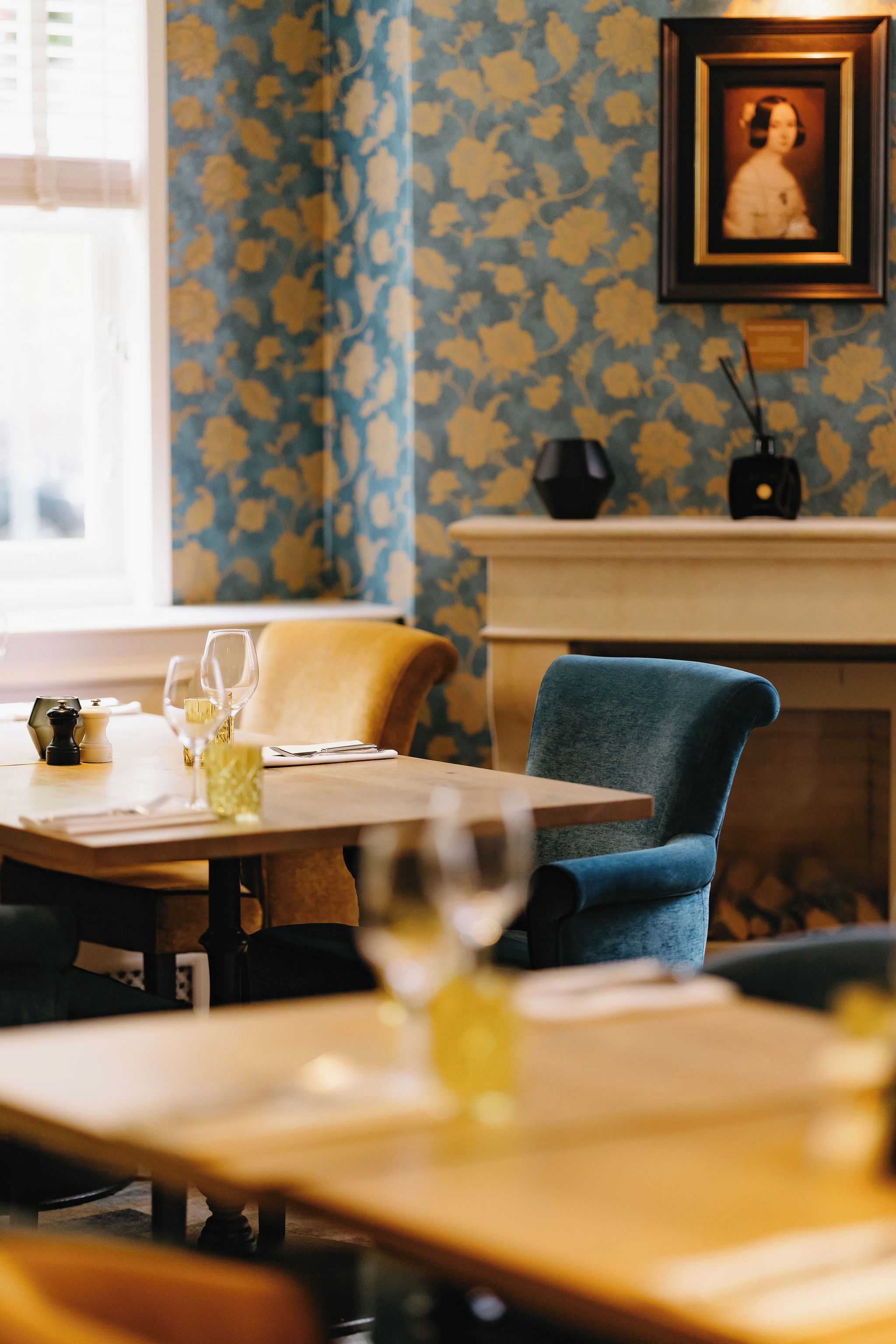 A dining area with a table set for two, a blue chair, and a fireplace in the background.