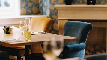 A dining area with a table set for two, a blue chair, and a fireplace in the background.