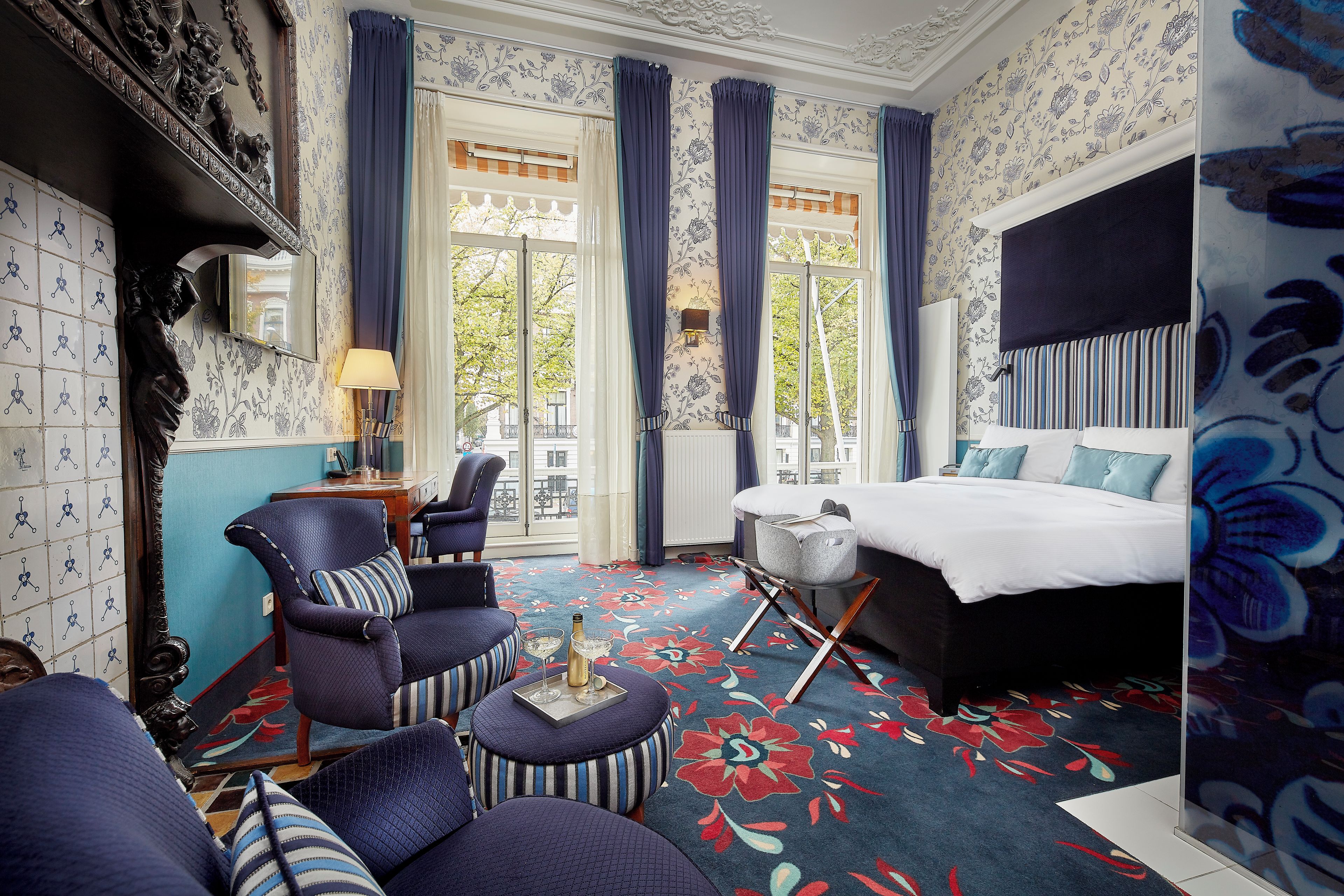 Habitación de hotel con una cama, dos sillones, una mesita con bebidas y vistas a la calle a través de grandes ventanales.