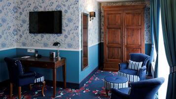 A room with a blue and white floral wallpaper, a wooden wardrobe, a desk with a chair, a television mounted on the wall, and a window with curtains.