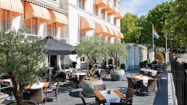 An outdoor cafe with tables and chairs, potted plants, and a building with striped awnings in the background. - Carlton Ambassador Hotel (The Hague)