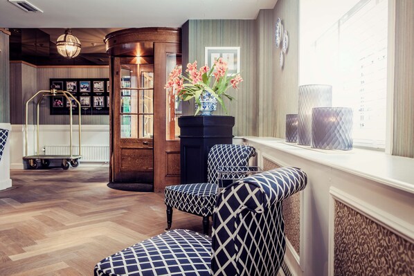A hotel lobby with patterned chairs, a vase with flowers, and a view of the city through large windows. - Carlton Ambassador Hotel (The Hague)
