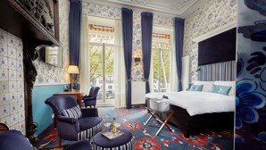 A hotel room with a large bed, a sitting area with a chair and ottoman, a small table with a bottle and glasses, and a view of the outdoors through a large window. - Carlton Ambassador Hotel (The Hague)