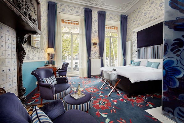 A hotel room with a large bed, a sitting area with a chair and ottoman, a small table with a bottle and glasses, and a view of the outdoors through a large window. - Carlton Ambassador Hotel (The Hague)