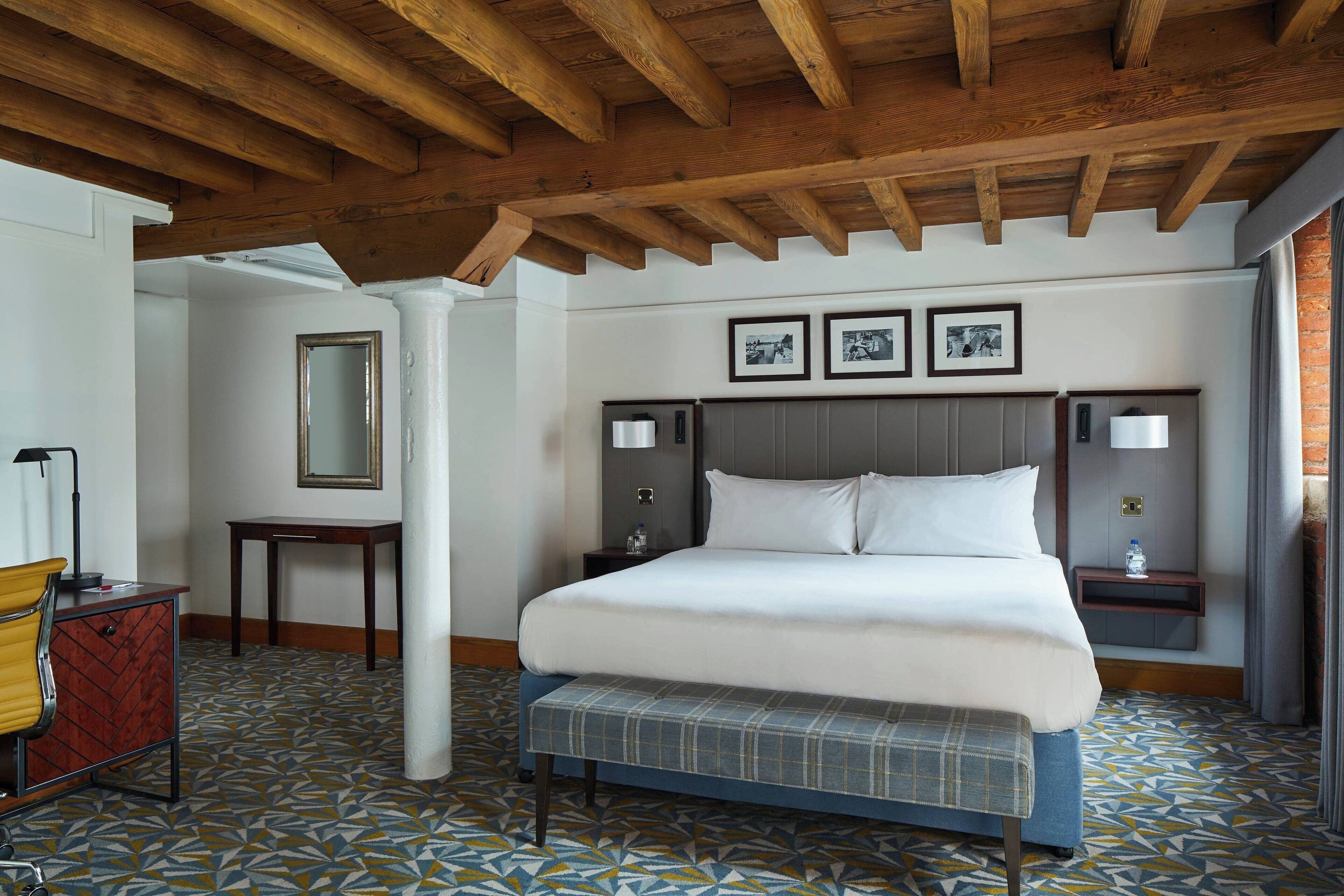 A hotel room with a large bed, a bench, a desk, and a mirror.