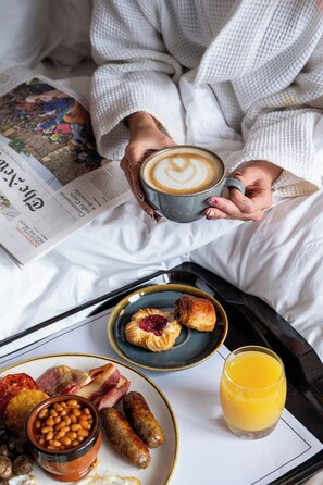 A person in a white robe holding a cup of coffee and a newspaper, with breakfast items on a tray. - Manchester Marriott Victoria & Albert Hotel (Manchester)