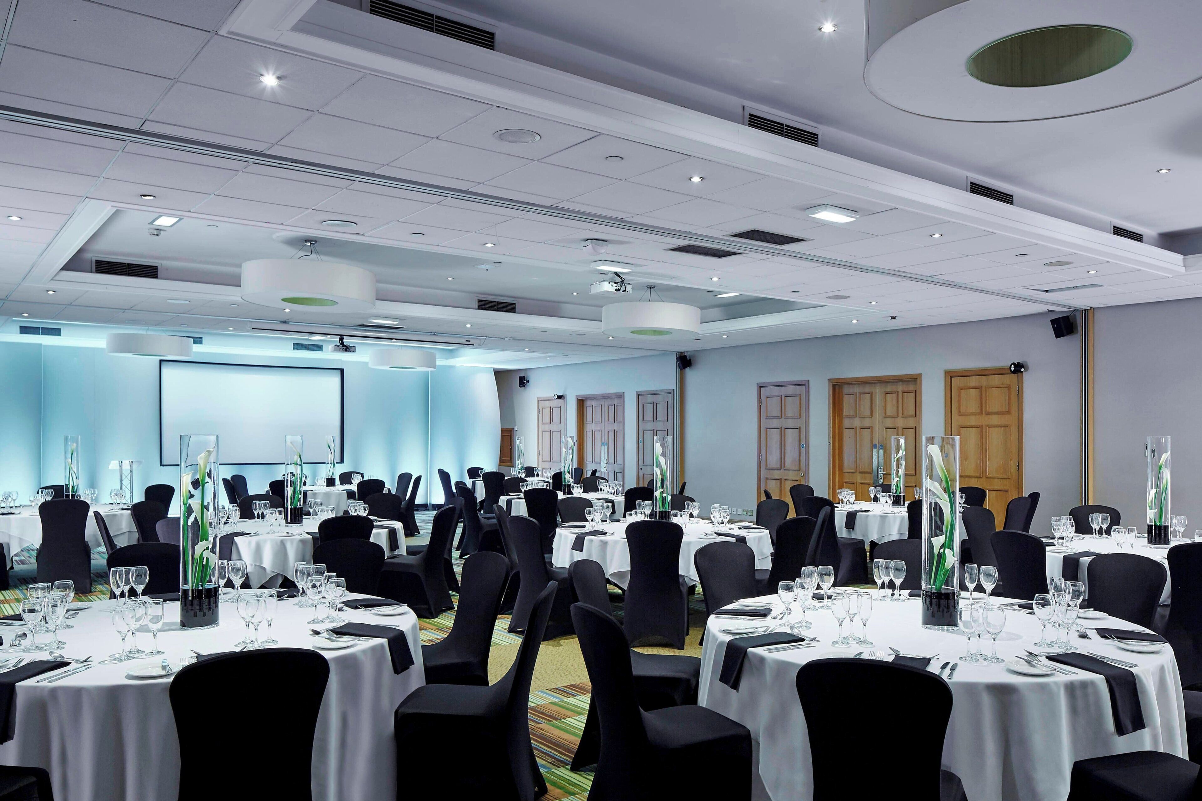 A conference room with multiple round tables, black chairs, and white tablecloths.