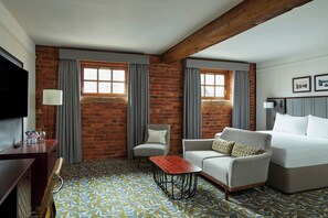 Una habitación de hotel con pared de ladrillo, dos ventanas con cortinas, un televisor de pantalla plana, un sofá, una mesa de centro y una cama.