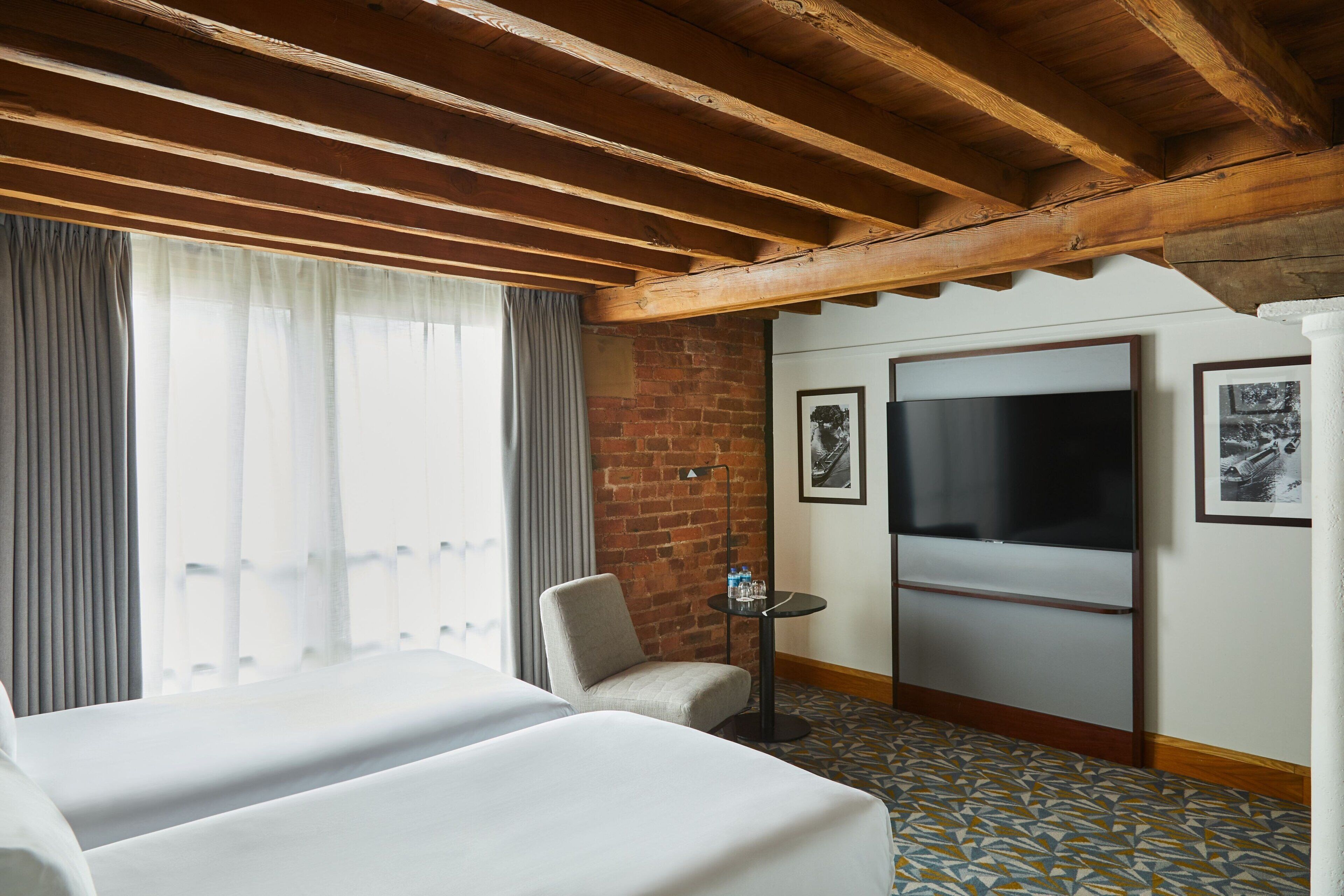 Een hotelkamer met een bed, een stoel, een tafel met waterflesjes, een televisie en een bakstenen muur.