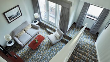 A hotel room with a sofa, armchair, and a red ottoman. There is a large window with curtains, a framed picture on the wall, and a patterned floor.
