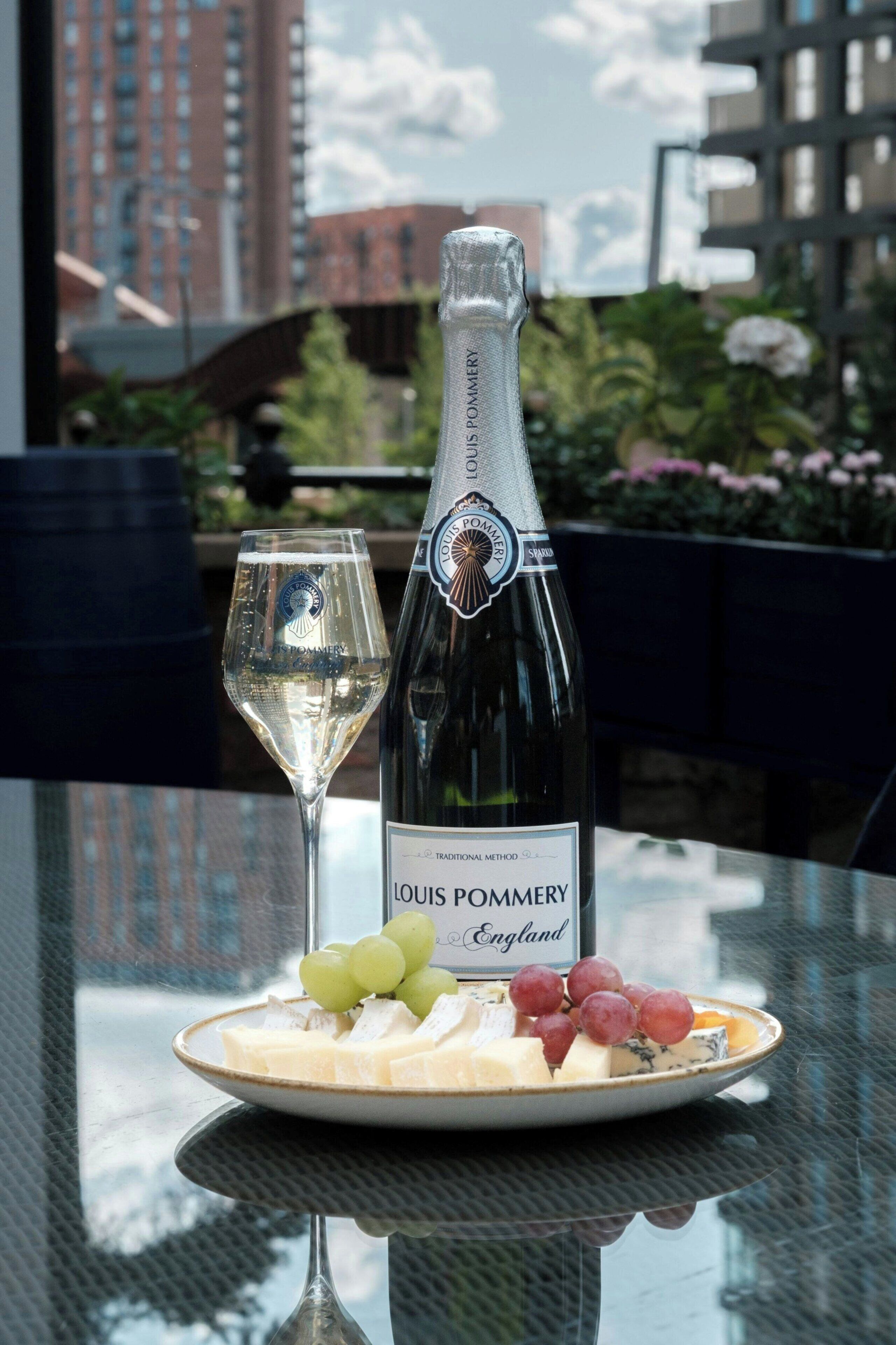 Una botella de champán Louis Pommery, una copa de champán y un plato con queso y uvas sobre una mesa con vista a la ciudad.