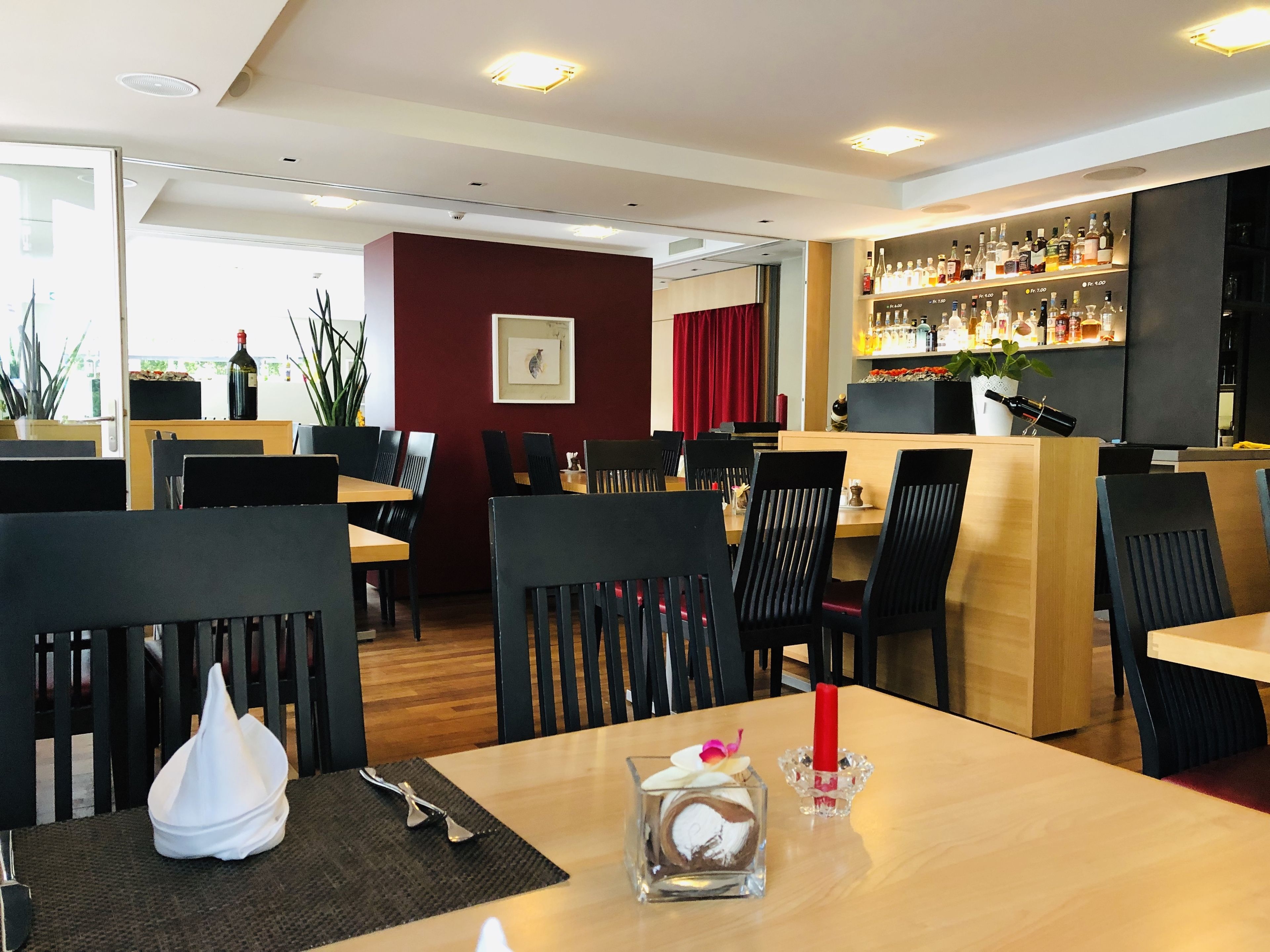Un restaurant avec des tables en bois et des chaises noires, un coin bar avec des bouteilles d’alcool exposées, et un rideau rouge en arrière-plan.