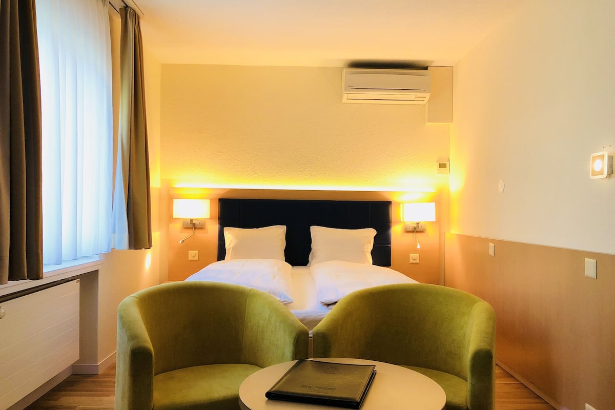 A hotel room with a bed, two green armchairs, a small round table, and a window with curtains.