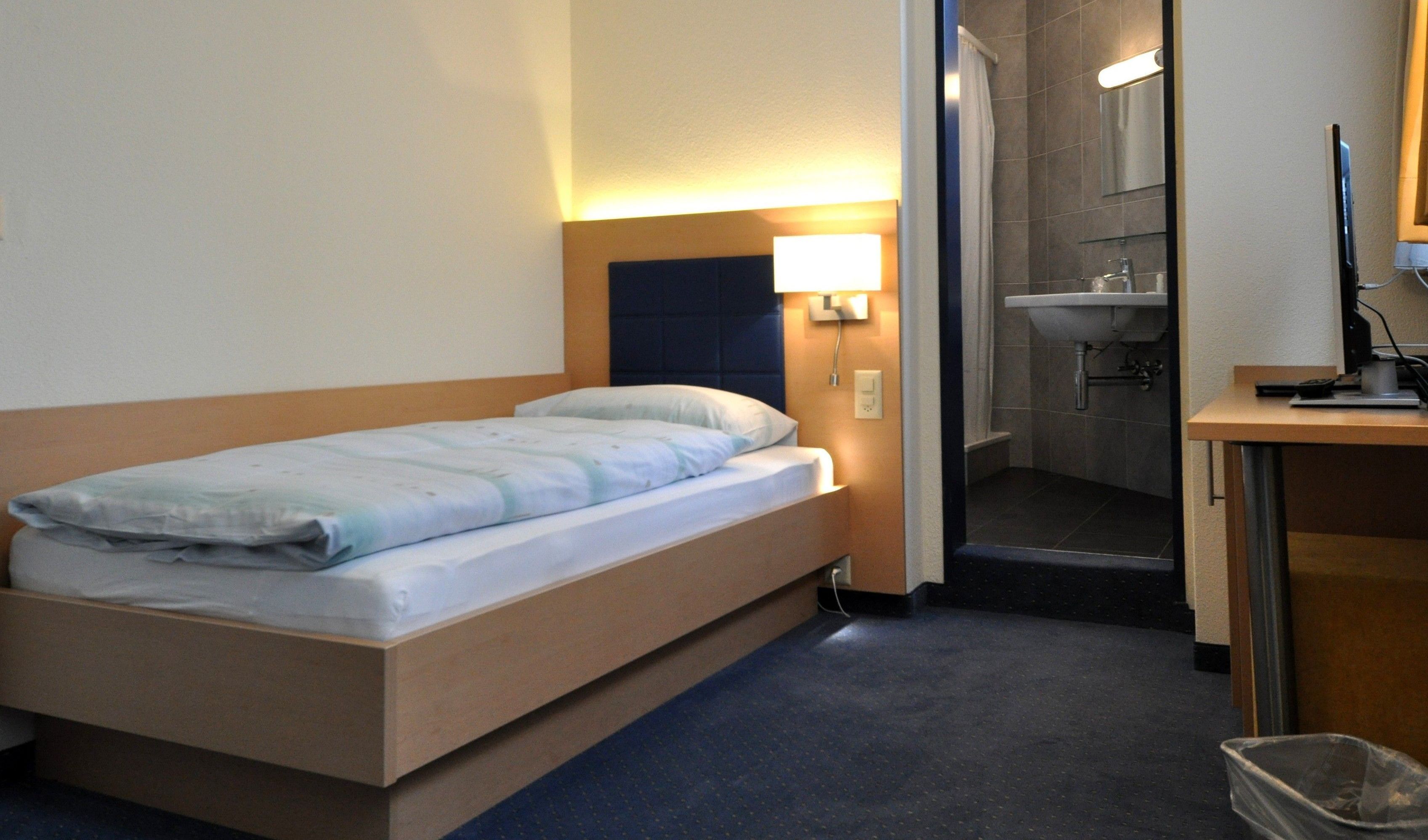 Habitación de hotel con una cama individual, una mesita de noche, un escritorio con computadora y un baño con lavamanos y espejo.