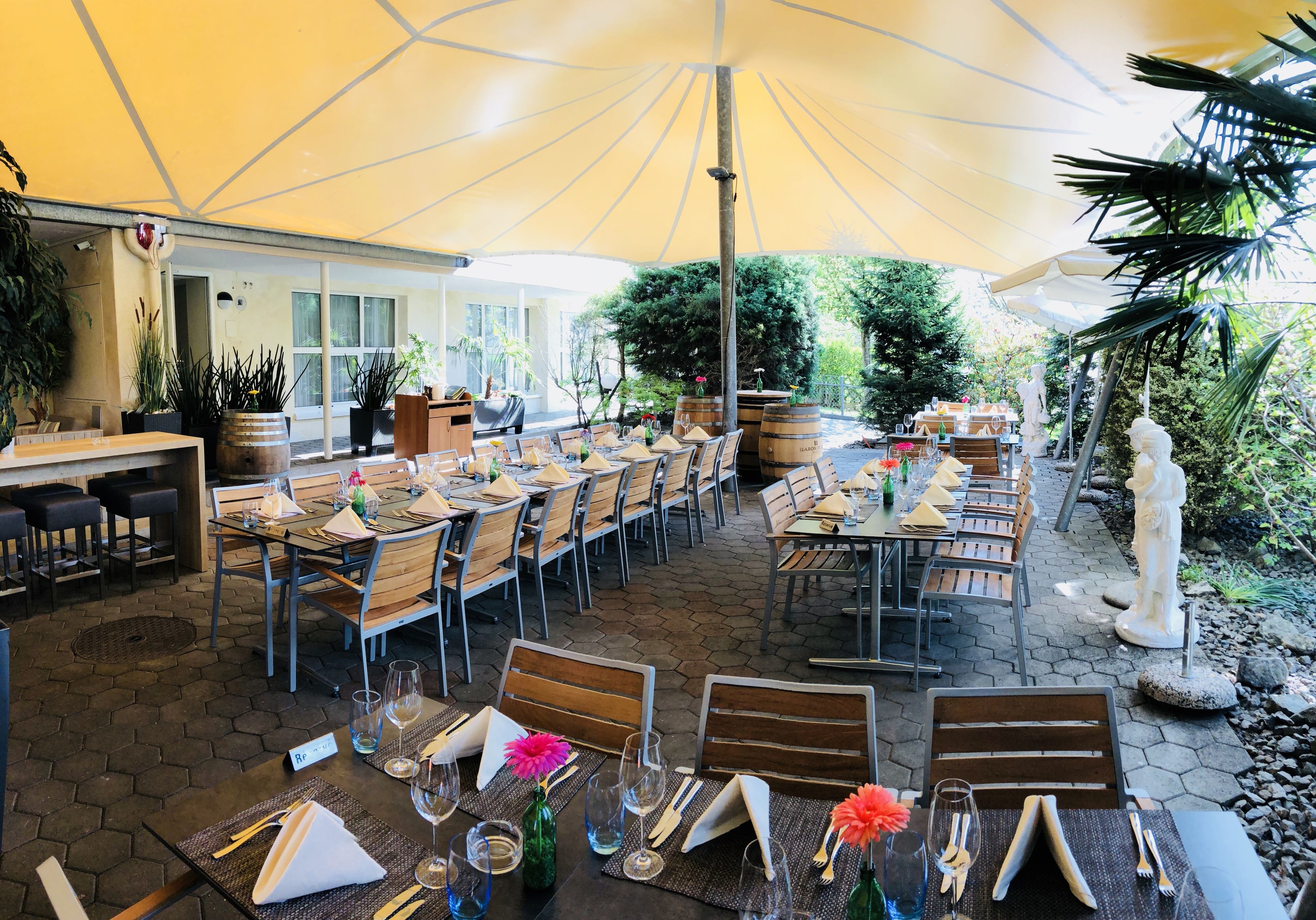 Un espace de restauration en plein air, doté de plusieurs tables et chaises, entouré de plantes en pot et d’une statue.