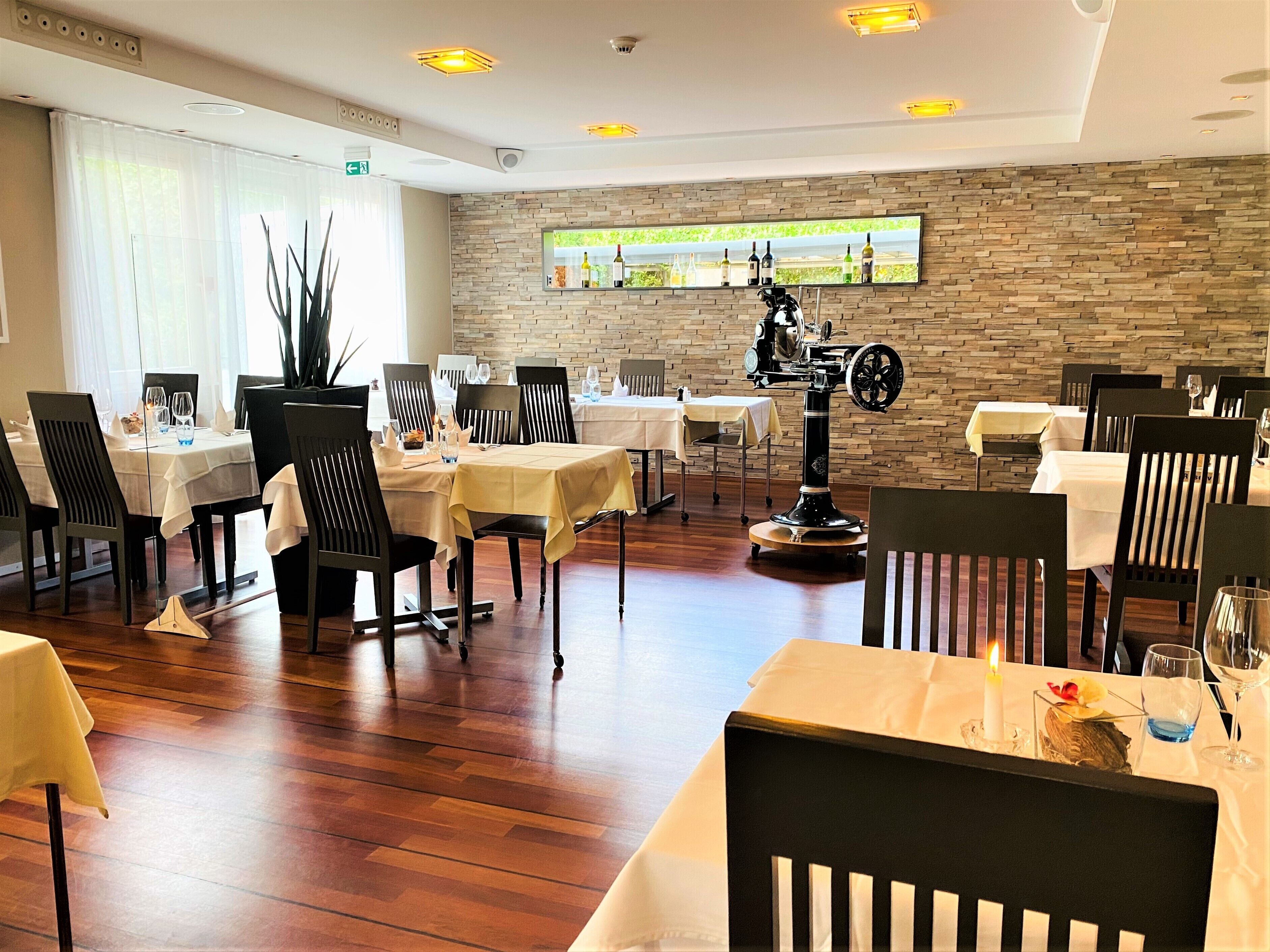 Un restaurante con suelo de madera, mesas con manteles blancos, sillas negras y una pared de piedra con una estantería para vinos.