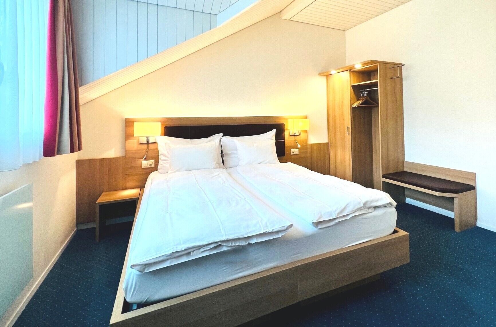 Quarto de hotel moderno com uma cama grande, mesas de cabeceira, guarda-roupa e um banco.