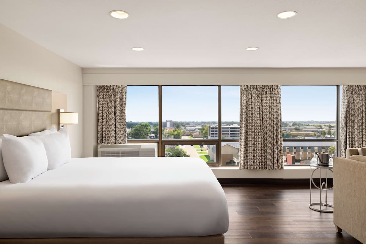 Un cuarto de hotel con una cama grande, un sofá, una mesita y vista a la ciudad a través de amplios ventanales.