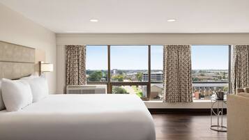 A hotel room with a large bed, a sofa, a small table, and a view of the city through large windows.