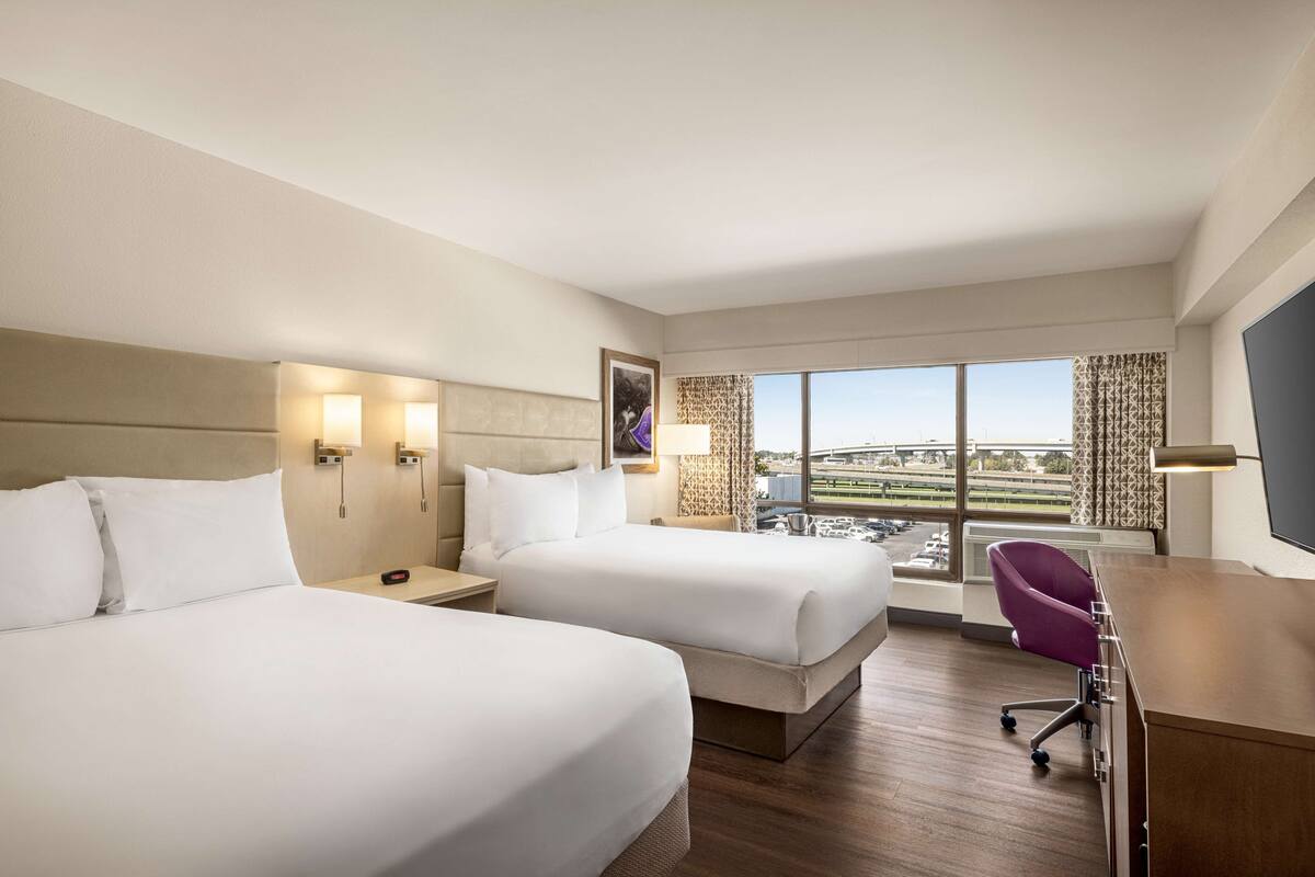 Habitación de hotel con dos camas, un escritorio, una silla y un amplio ventanal con vistas a la ciudad.