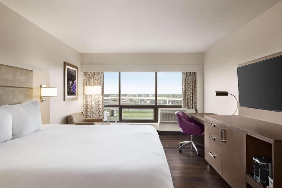 Habitación de hotel con una cama grande, un escritorio con silla, televisión y vista a la ciudad a través de la ventana.