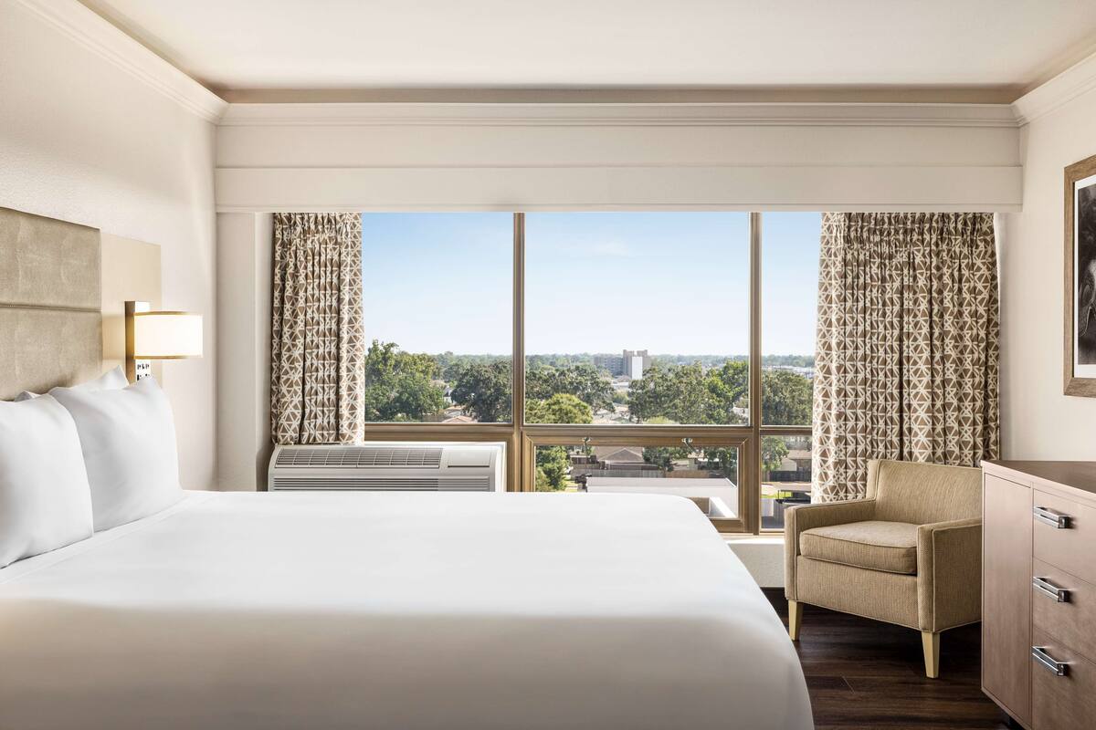 Habitación de hotel con una cama grande, una silla, un escritorio y vistas a la ciudad a través de amplios ventanales.