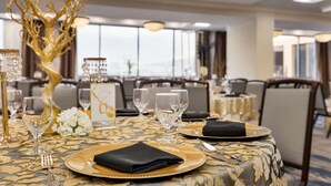 Una mesa de comedor decorada con vajilla dorada y negra, flores blancas y un centro de mesa con forma de árbol dorado, en una habitación luminosa con grandes ventanales.