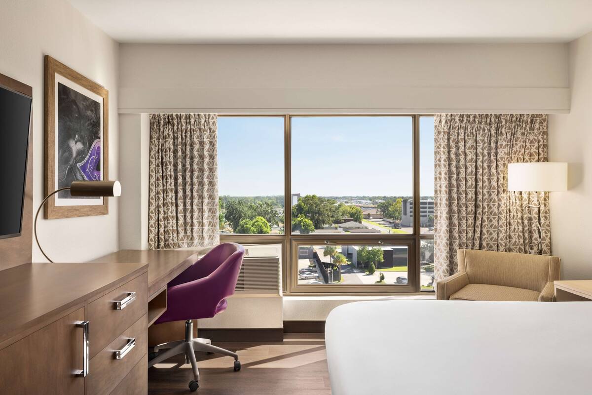 Una habitación de hotel con una cama, un escritorio con silla, un televisor y un amplio ventanal con cortinas.