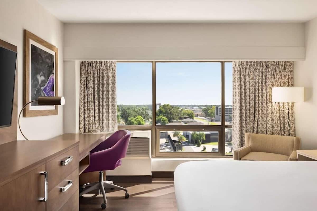 Una habitación de hotel con cama, escritorio, silla, televisor y un amplio ventanal con cortinas.