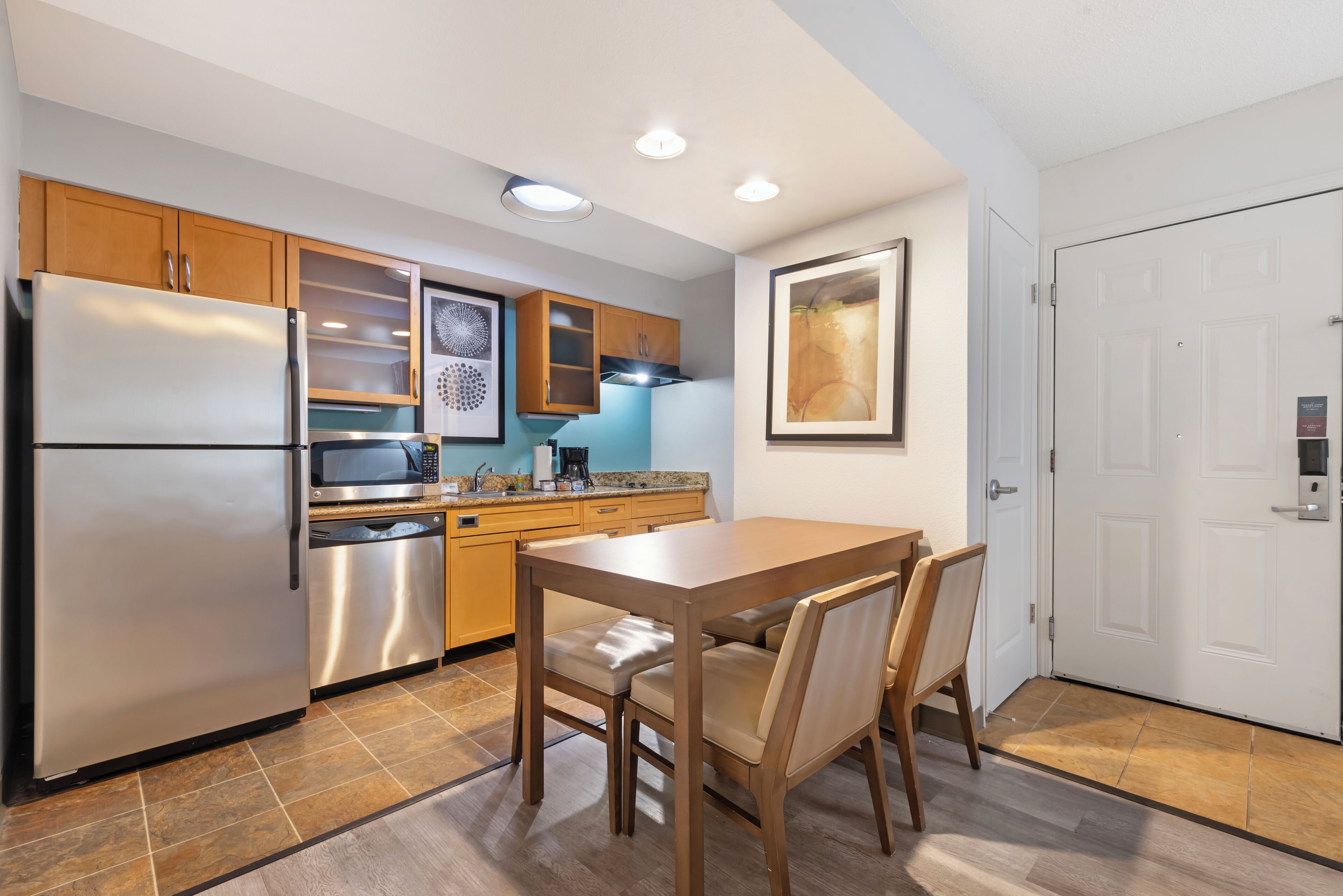 Cocina moderna con refrigerador de acero inoxidable, microondas y lavavajillas, armarios de madera y una mesa de comedor con sillas.