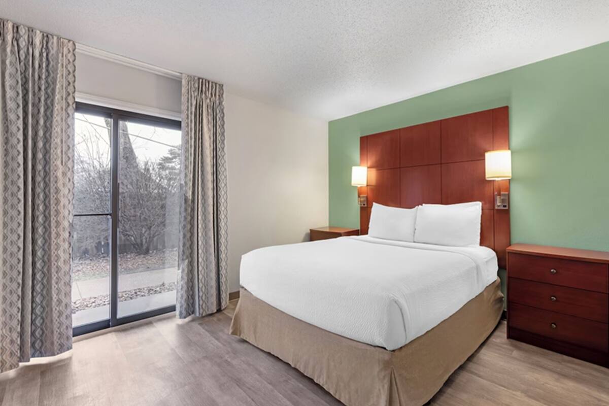 A hotel room with a bed, a nightstand, a lamp, a dresser, and a view through a glass door.
