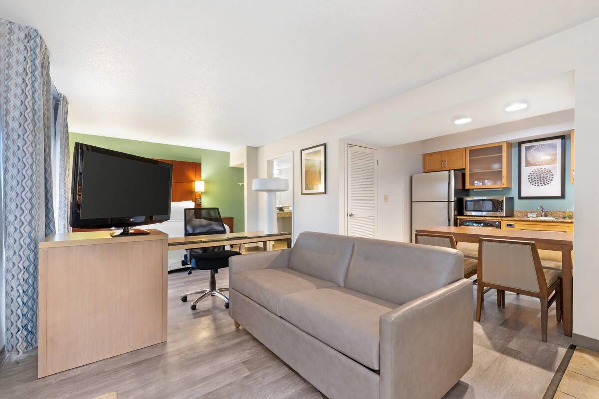 A modern hotel room with a sofa, TV, desk, and kitchenette.