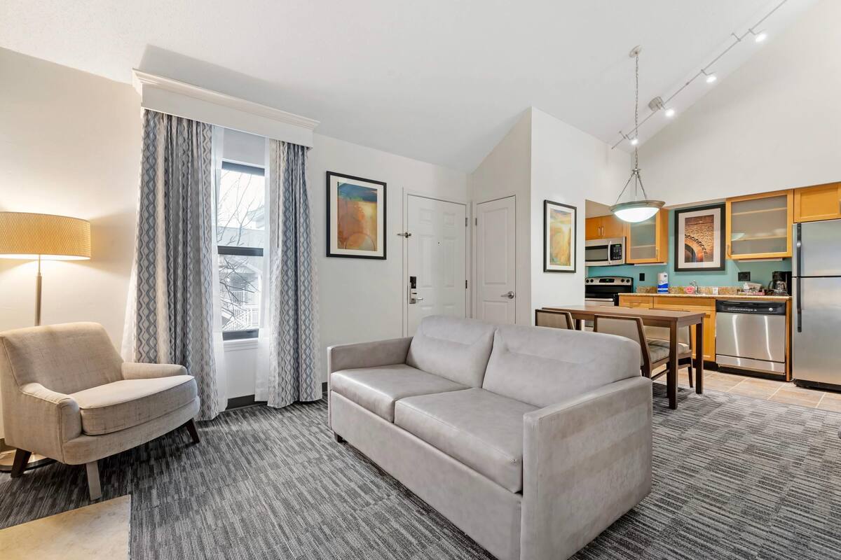 A modern living room with a sofa, armchair, and dining table. There is a kitchen area with appliances and a dining table.