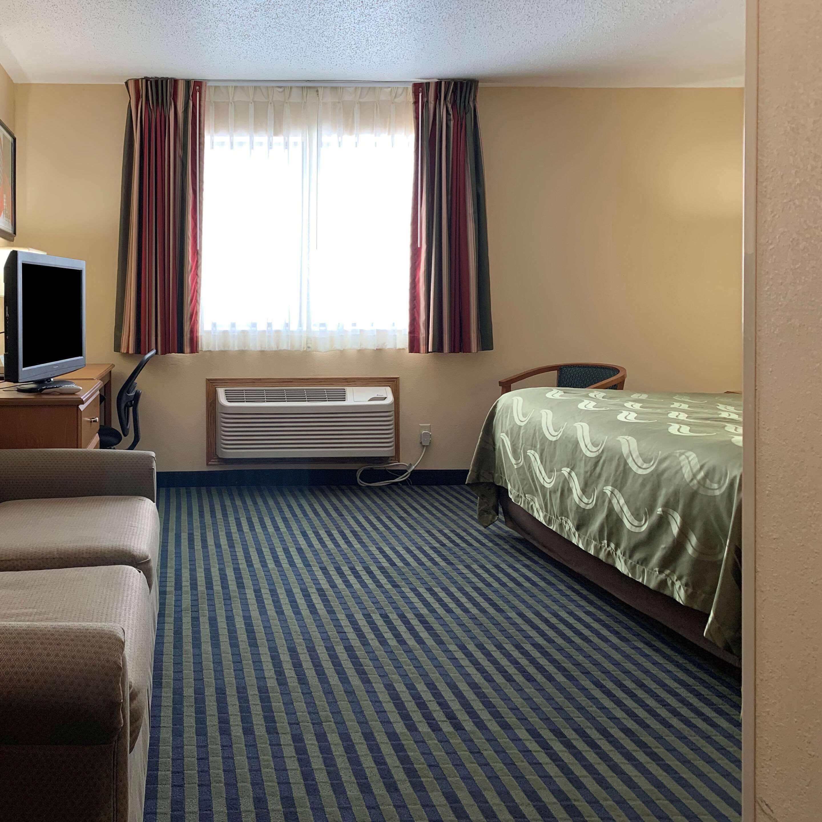 Une chambre d’hôtel avec un lit, un bureau équipé d’un ordinateur, un climatiseur et une fenêtre avec des rideaux.