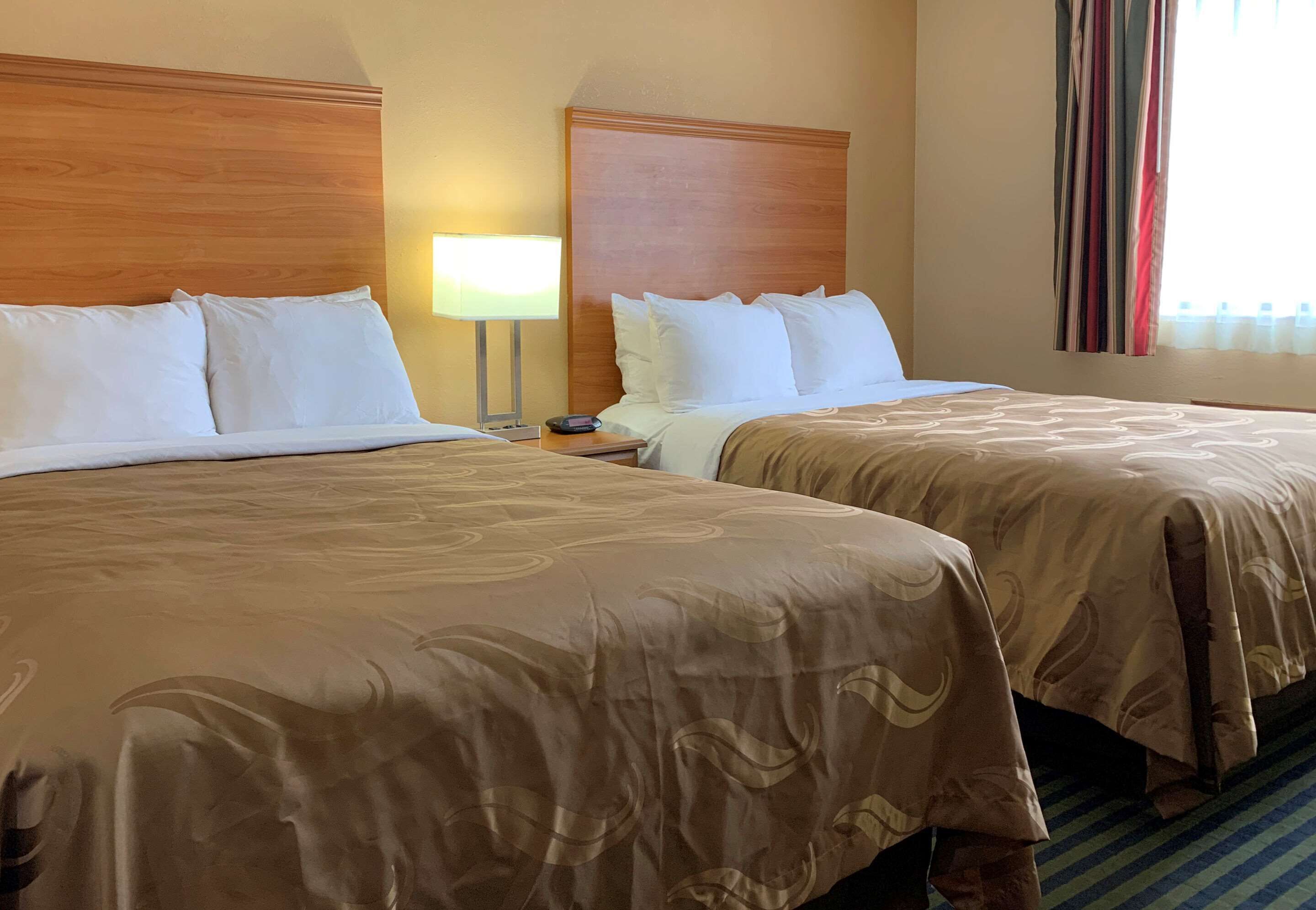 A hotel room with two beds, each with white linens and a brown bedspread. There is a nightstand with a lamp between the beds.