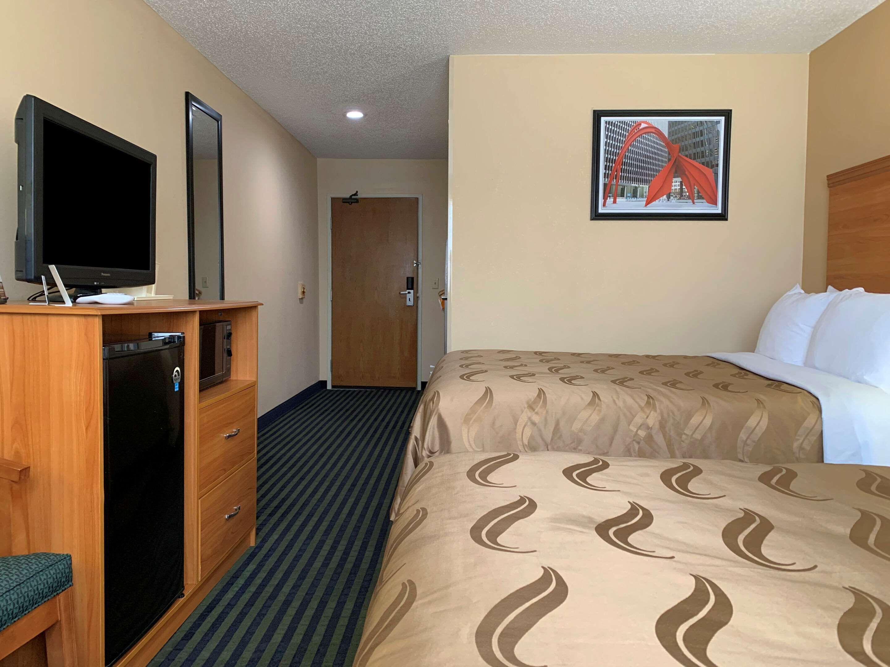 A hotel room with a bed, a desk with a television, a refrigerator, a door, and a framed picture on the wall.