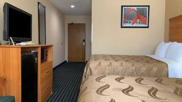 A hotel room with a bed, a desk with a television, a refrigerator, a door, and a framed picture on the wall.