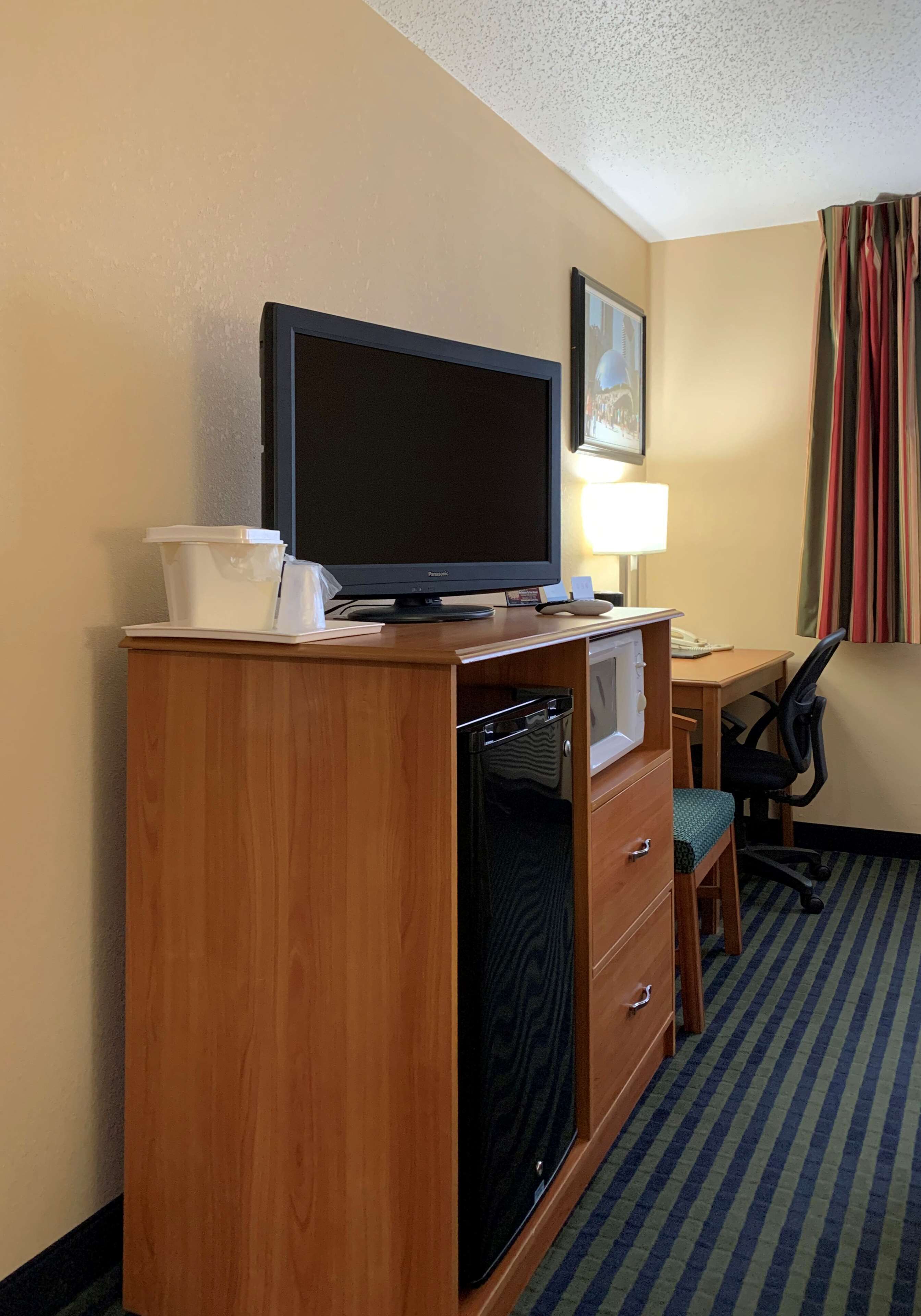 A hotel room with a wooden desk, a television, a microwave, a lamp, and a chair.