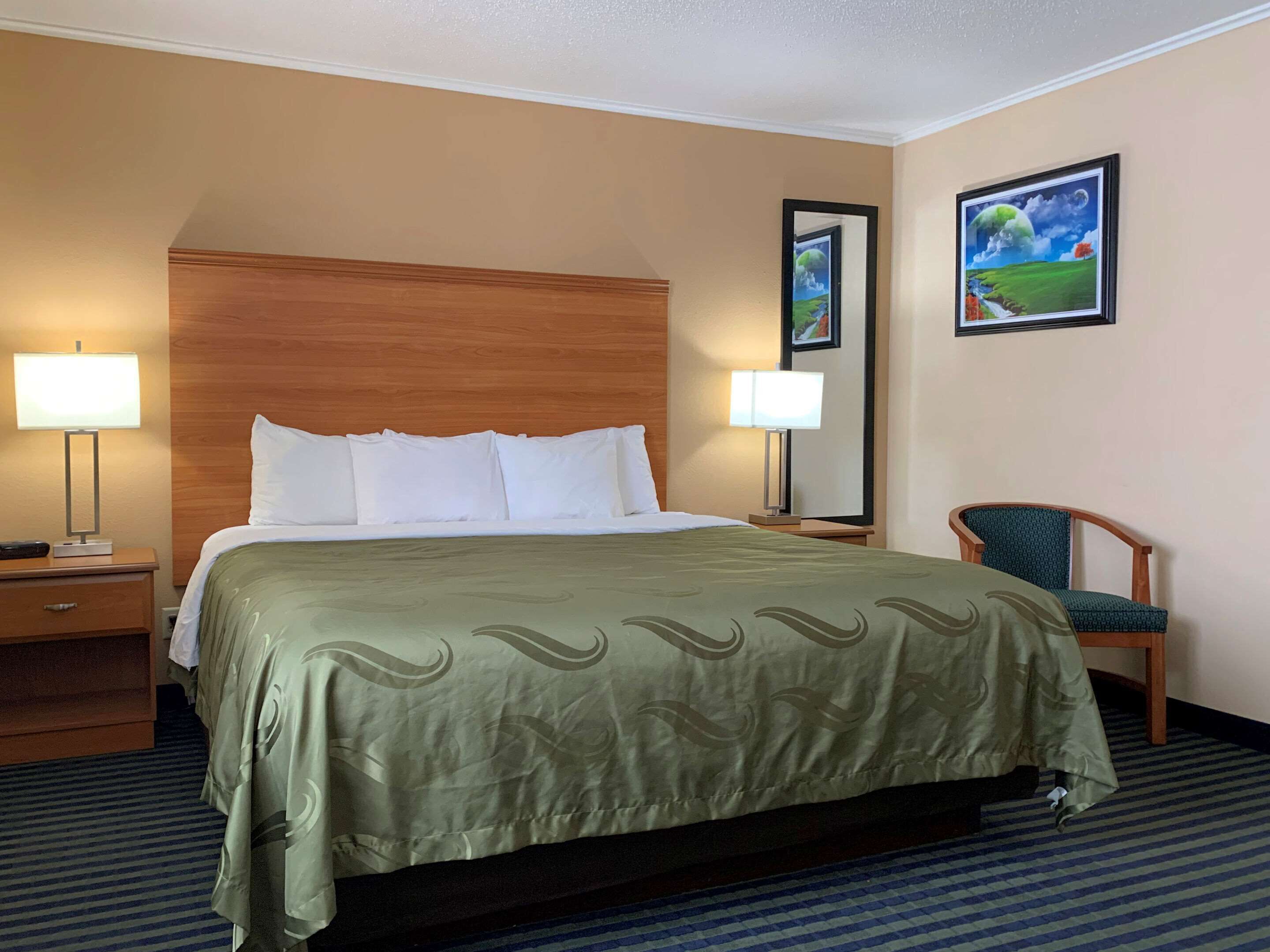 A hotel room with a large bed, two bedside lamps, a wooden headboard, a green patterned bedspread, a blue-striped carpet, a green chair, and two framed pictures on the wall.